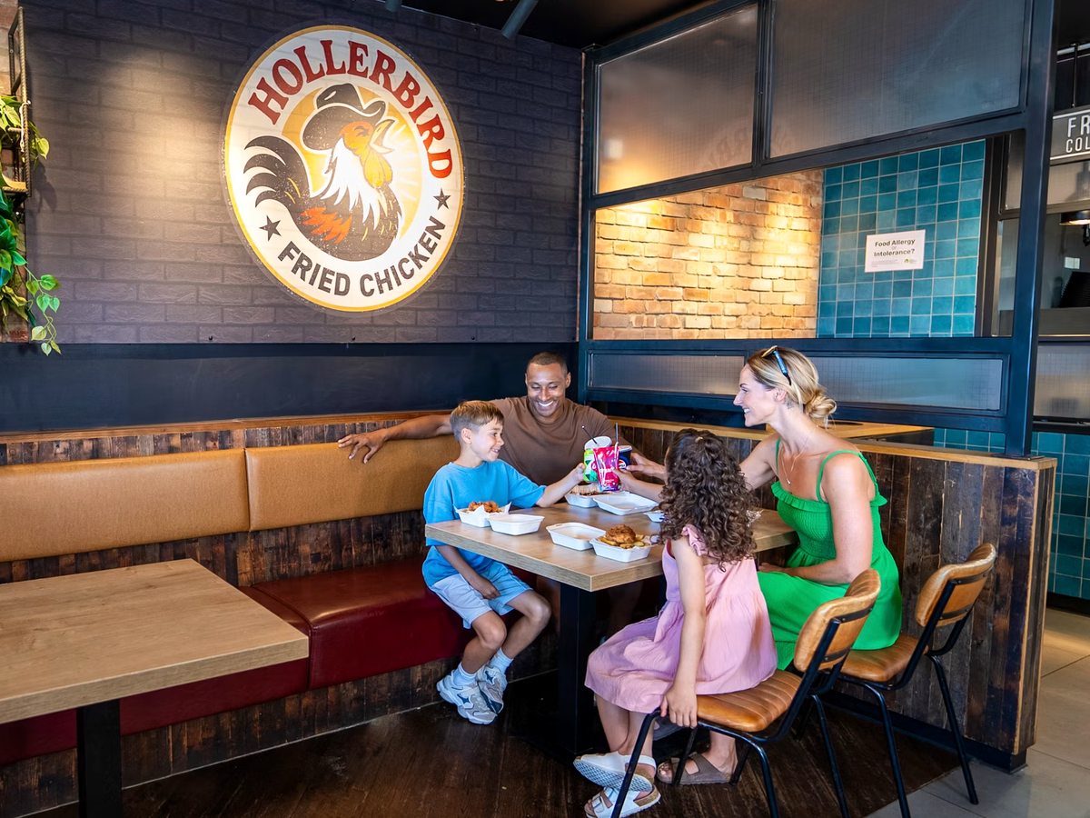 A family eating at a table in a padded booth at Hollerbird Fried Chicken, with the outlet's rooster logo and tiled walls behind.
