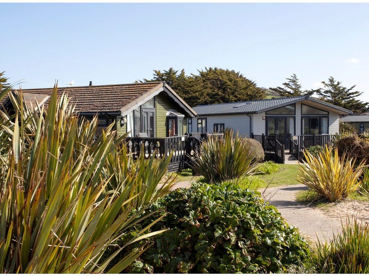 Two holiday lodges with pitched tiled roofs and dark timber verandas among coastal shrubs and spiky phormium planting, with a paved path running between them.