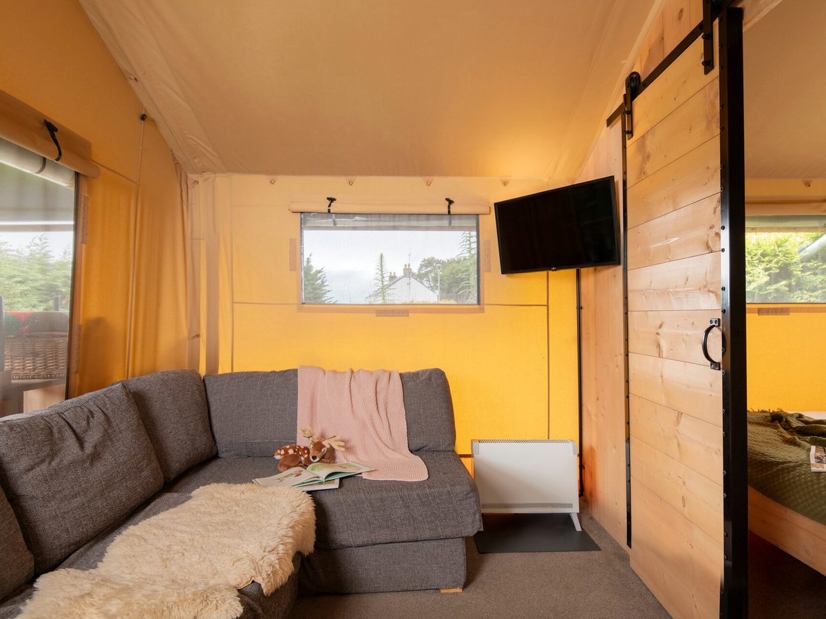 A safari tent living area with a grey corner sofa, a sheepskin throw and a wall-mounted television, with sliding timber doors through to the bedrooms.