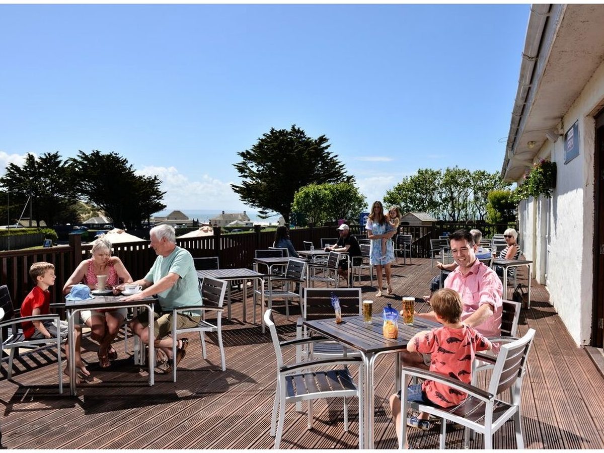 Families at metal tables and chairs across a wide timber terrace beside a white building, with trees and a strip of sea in the distance.