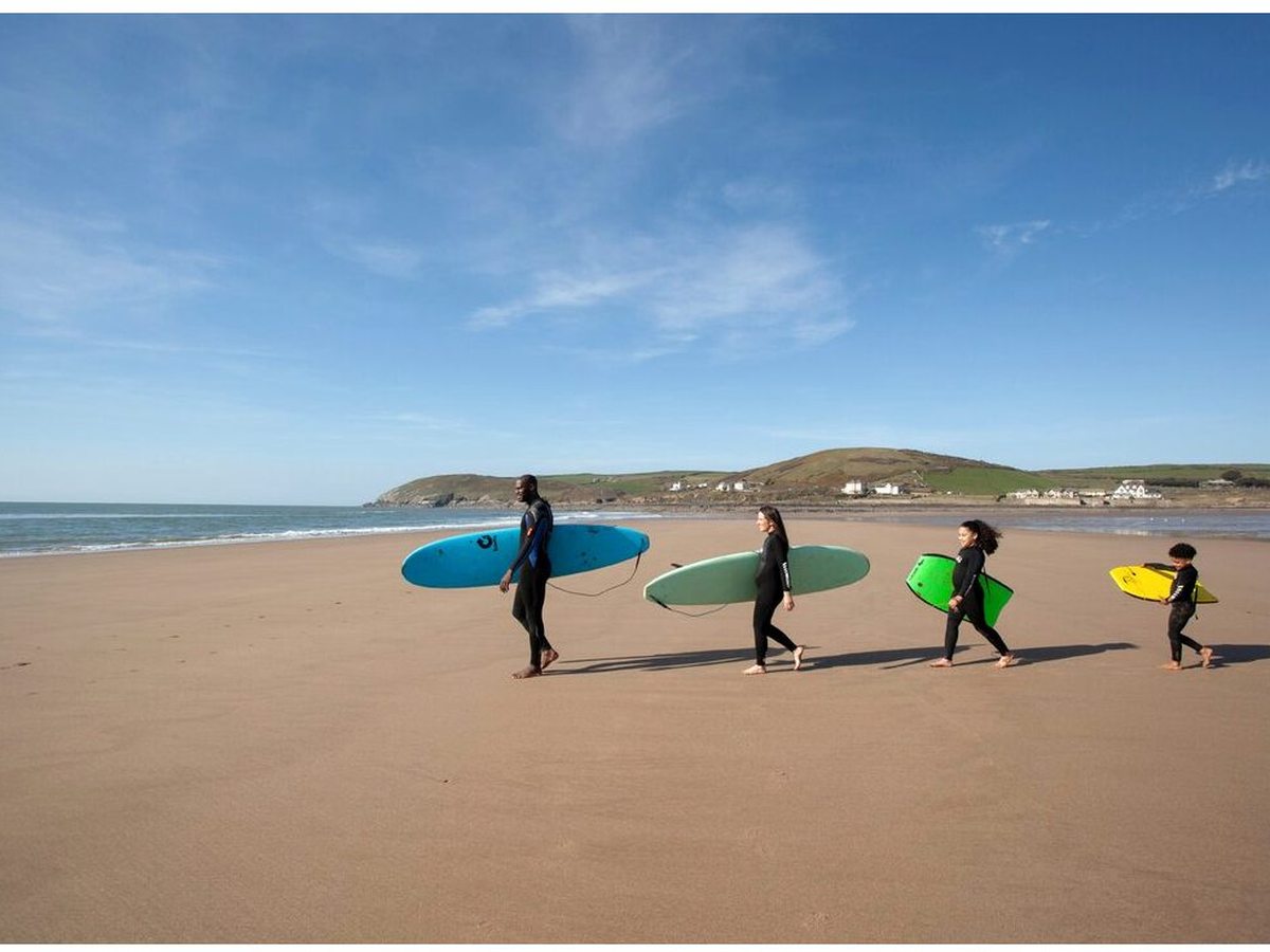 Four people in wetsuits carrying surfboards and bodyboards across firm wet sand, with the headland and village on the far side of the bay.