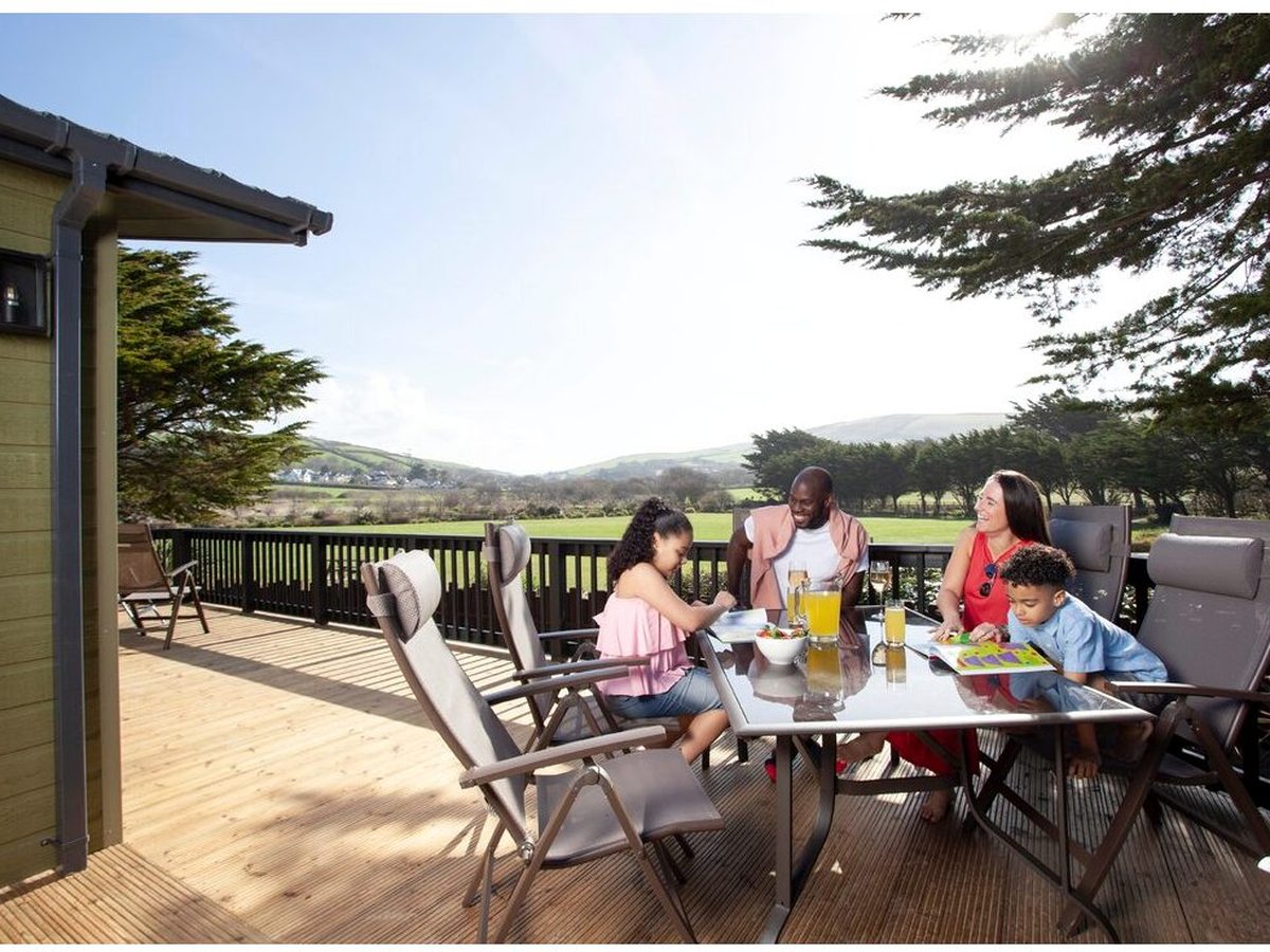 A family of four eating at a glass-topped table with cushioned recliners on a wide timber lodge veranda, with green fields and hills beyond the railing.