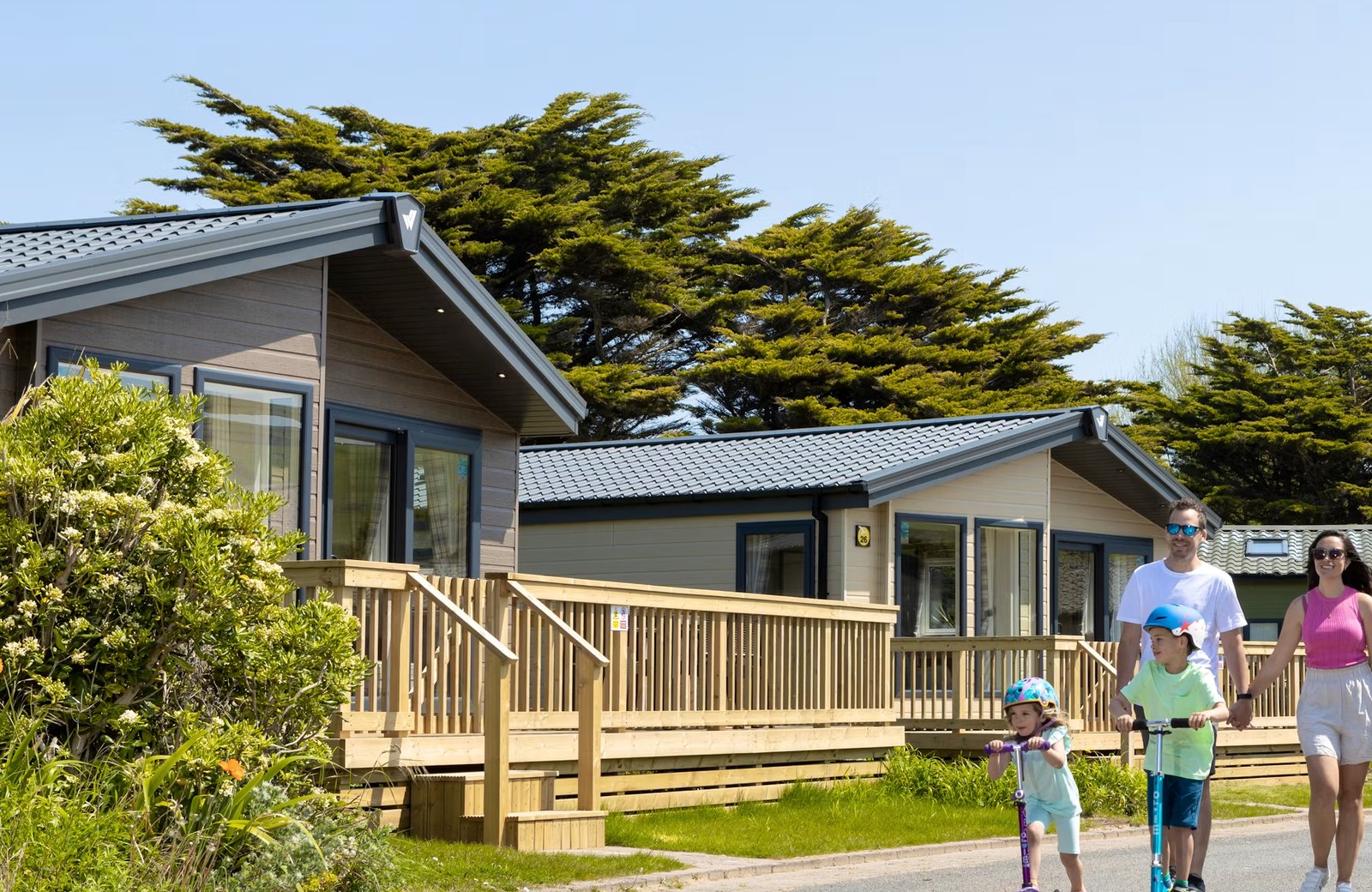 Two timber holiday lodges with tiled roofs and wooden verandas beside a paved road at Ruda Holiday Park, with a family walking and two children on scooters under a clear blue sky.