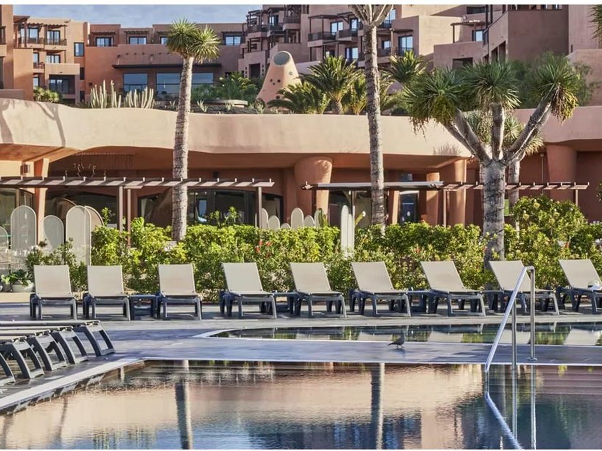 A long pool with a row of sun loungers along its paved edge, a clipped hedge and palms behind, and terracotta buildings stepping up the hillside beyond.