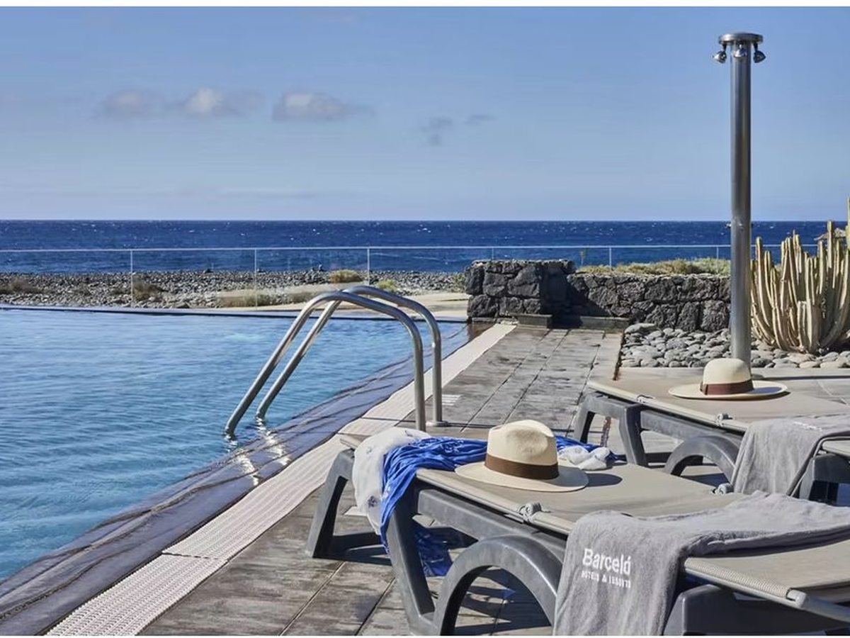 A pool at the top of a stony shoreline, with a metal ladder and handrail, a poolside shower, sun loungers with towels and a straw hat, and open blue sea beyond.