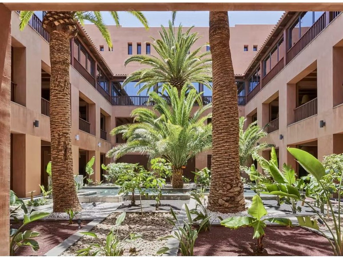 A courtyard between three-storey terracotta room blocks, planted with tall date palms and banana plants around a narrow water channel, with open arcaded walkways on each side.