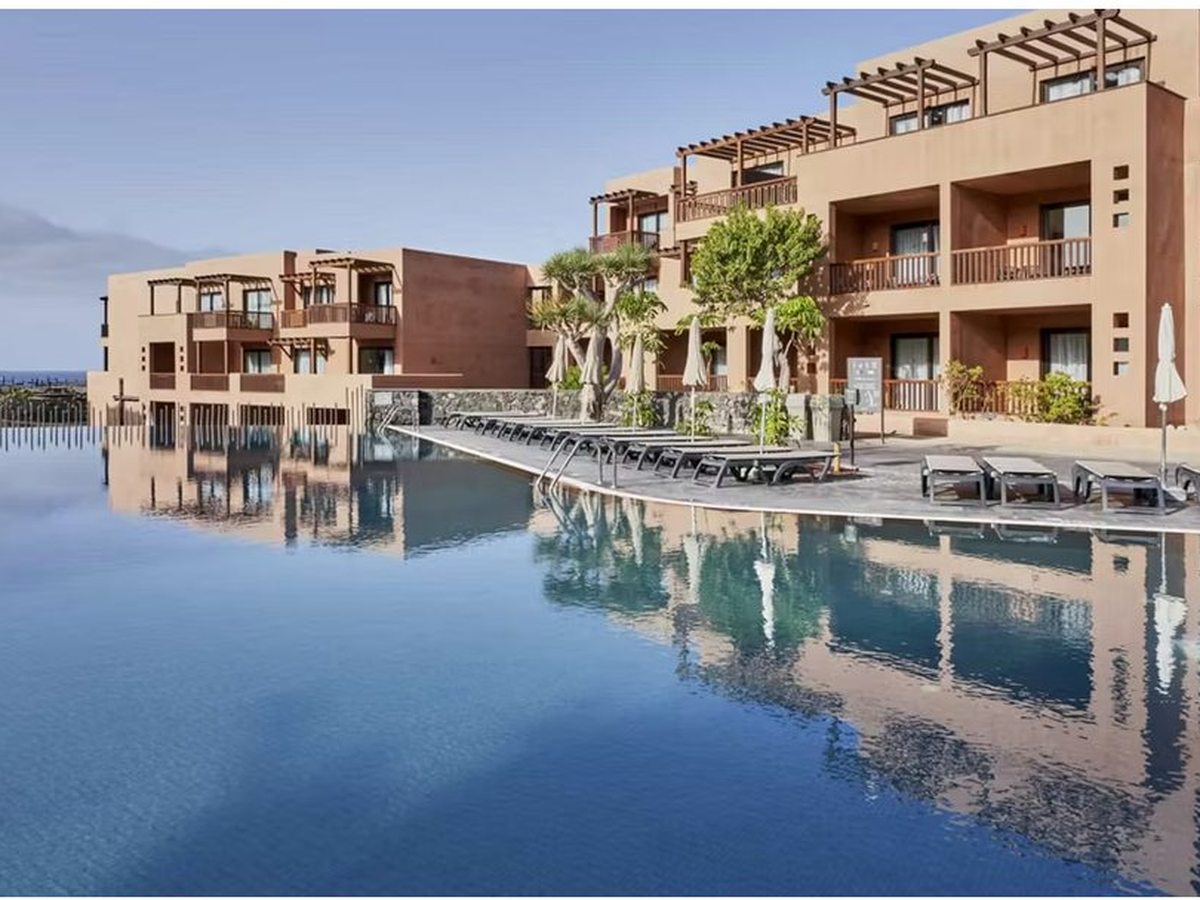 A large still pool reflecting the terracotta room blocks along its edge, with sun loungers, closed parasols and dragon trees on the paved surround.