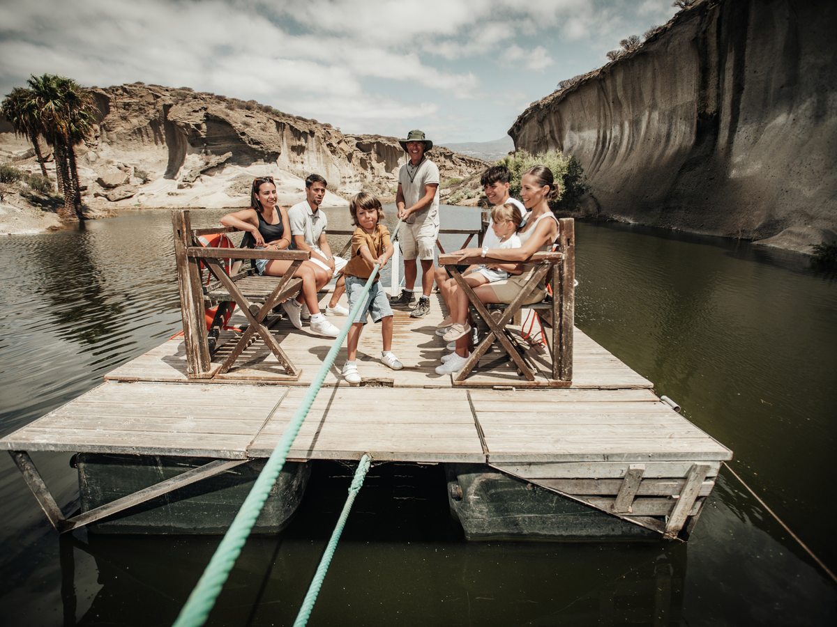 A family sitting on a timber raft on a still lake, pulled along by a rope, with eroded pale cliffs rising on both sides.