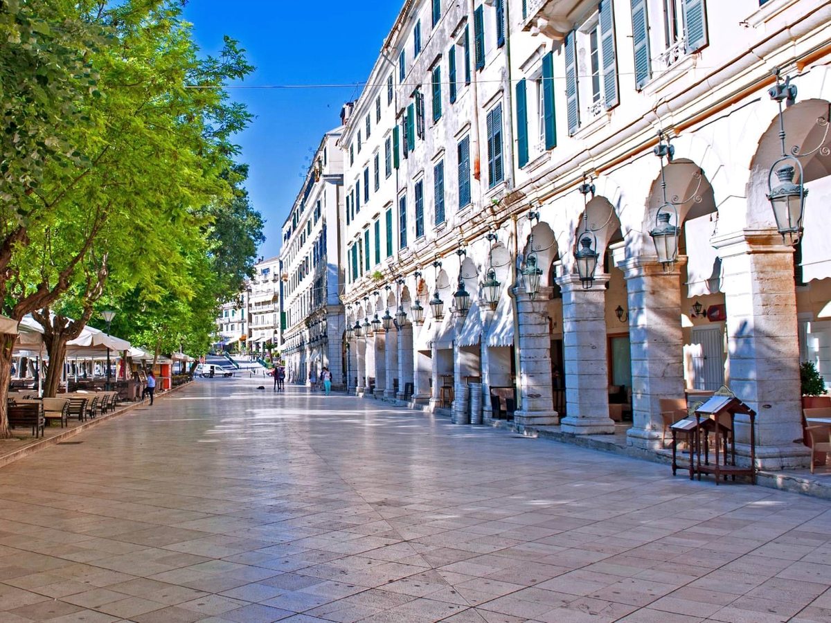A wide paved promenade beside a long arcade of stone arches hung with lanterns, with cafe tables under trees on one side and tall Venetian buildings above.