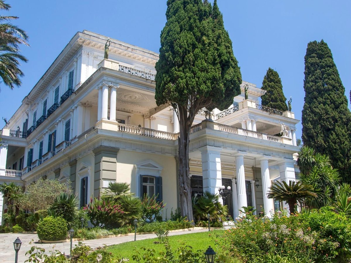 A neo-classical palace of pale stone with columned balconies and statues along its roofline, framed by tall cypresses and palms above terraced gardens.