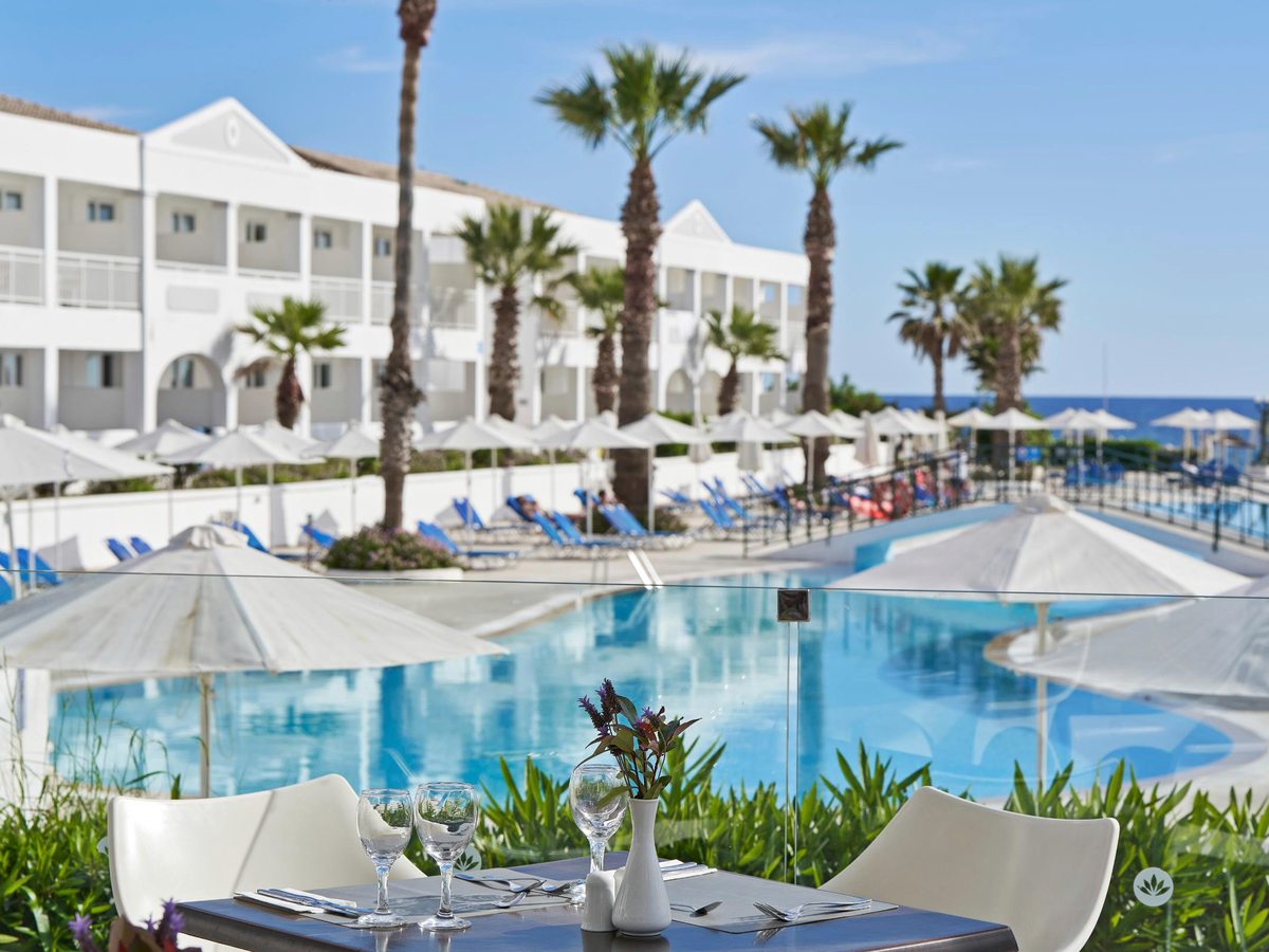 A long white two-storey accommodation block with arched balconies behind palms and rows of white parasols, above a large blue pool with blue loungers and the sea on the horizon.