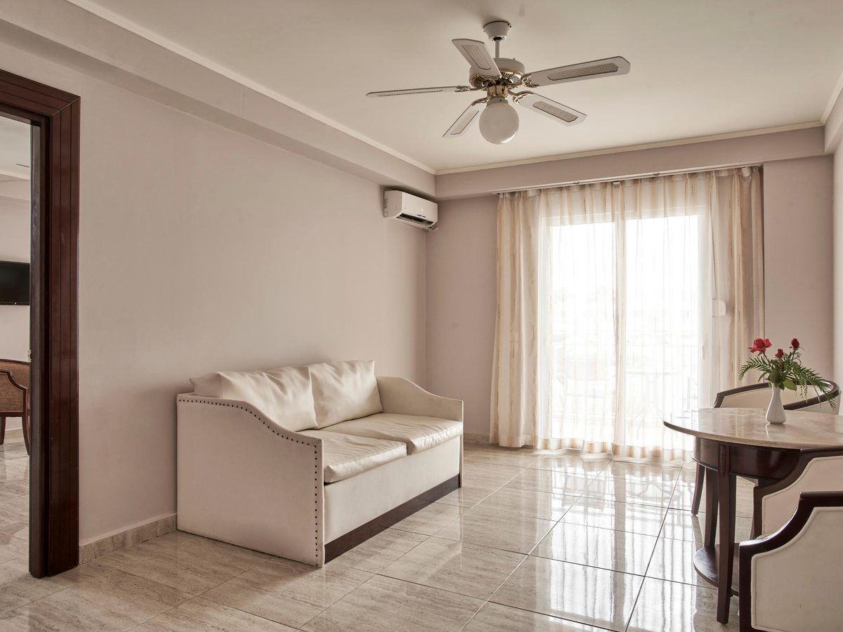 A suite sitting area with a cream sofa, a round dining table and chairs, a ceiling fan and full-length curtains at the balcony doors.
