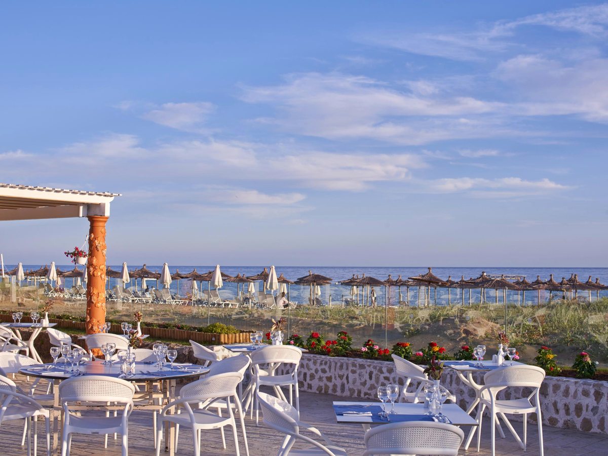 An open terrace of white tables and chairs behind a low stone wall, with rows of thatched parasols on the sand and the sea beyond.