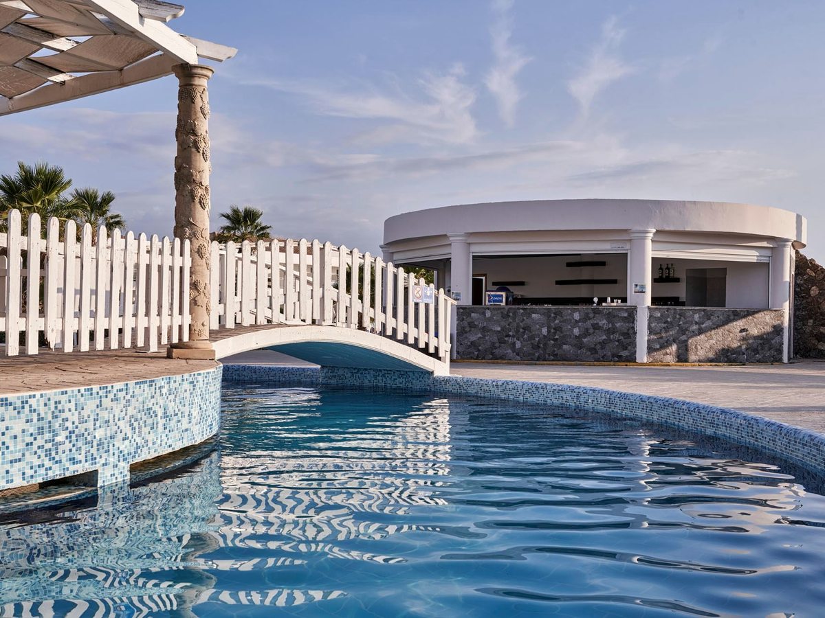 A curved pool edged with blue mosaic tiles, a white footbridge crossing it and a round open-fronted bar on the terrace beyond, with a white picket fence and a pergola.