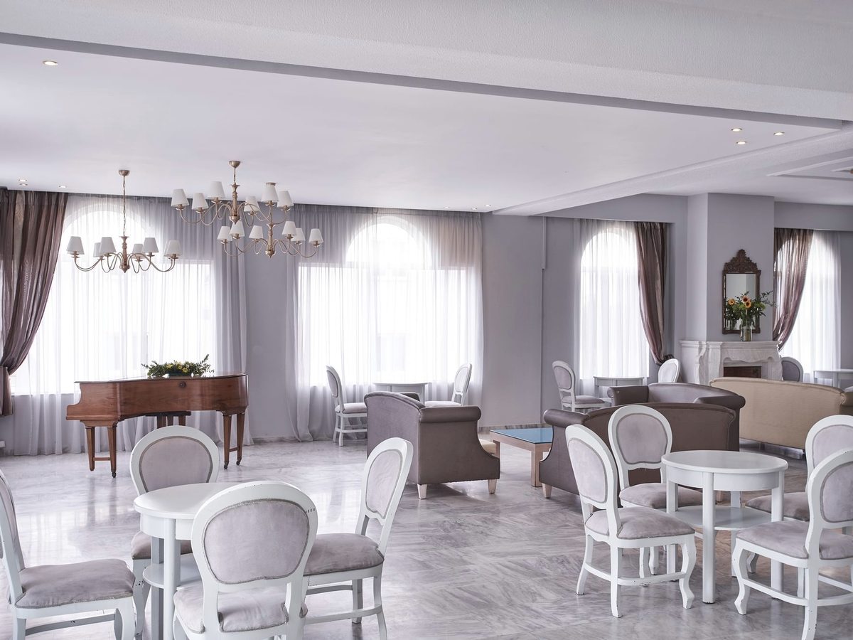 A large hotel lounge with white and grey armchairs around small round tables, a grand piano, chandeliers and tall arched windows hung with pale curtains.