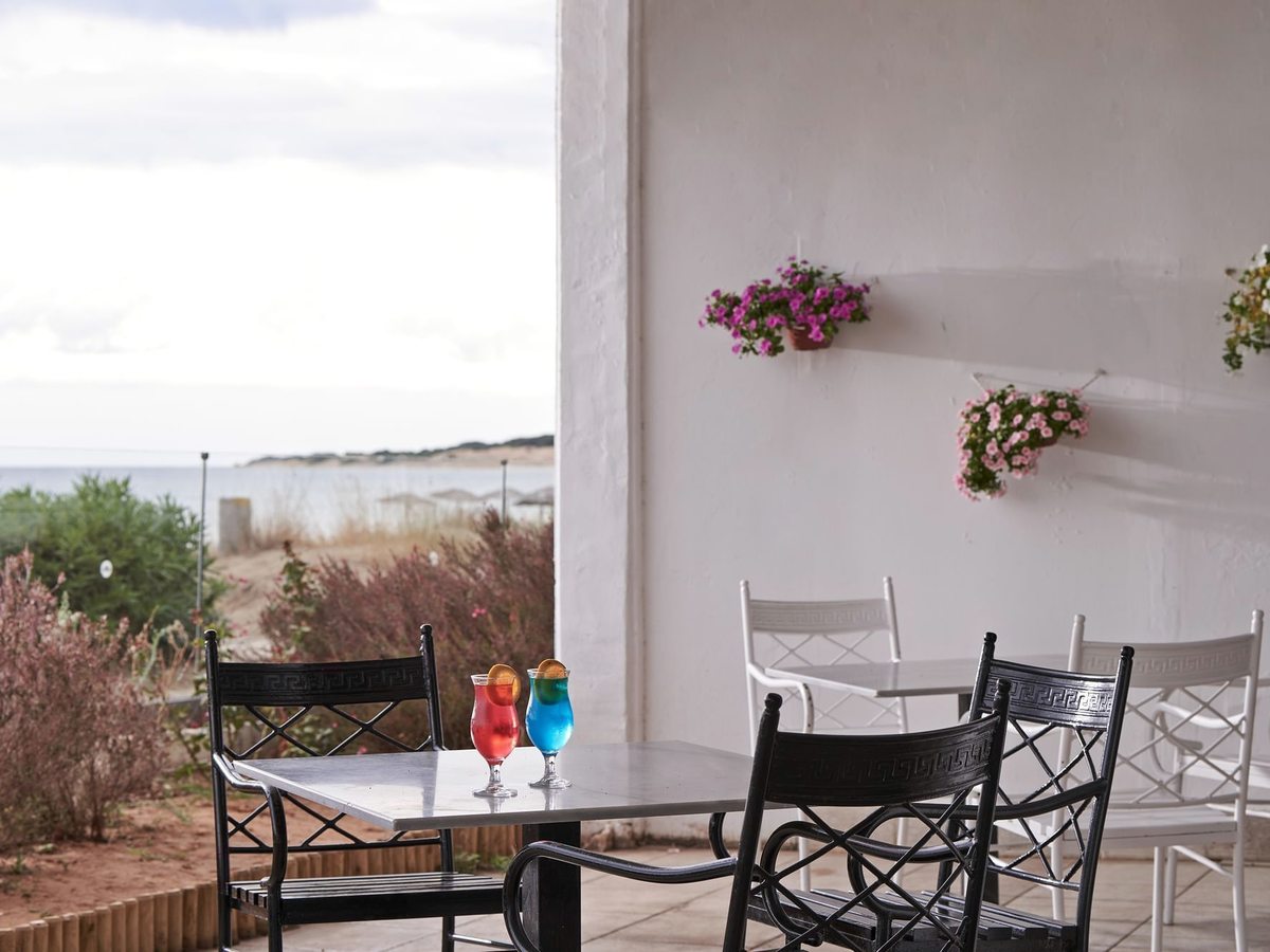 An open-sided bar terrace with black metal chairs at white tables, two tall coloured cocktails on a table, hanging flower baskets on a white wall and dunes and the sea beyond.