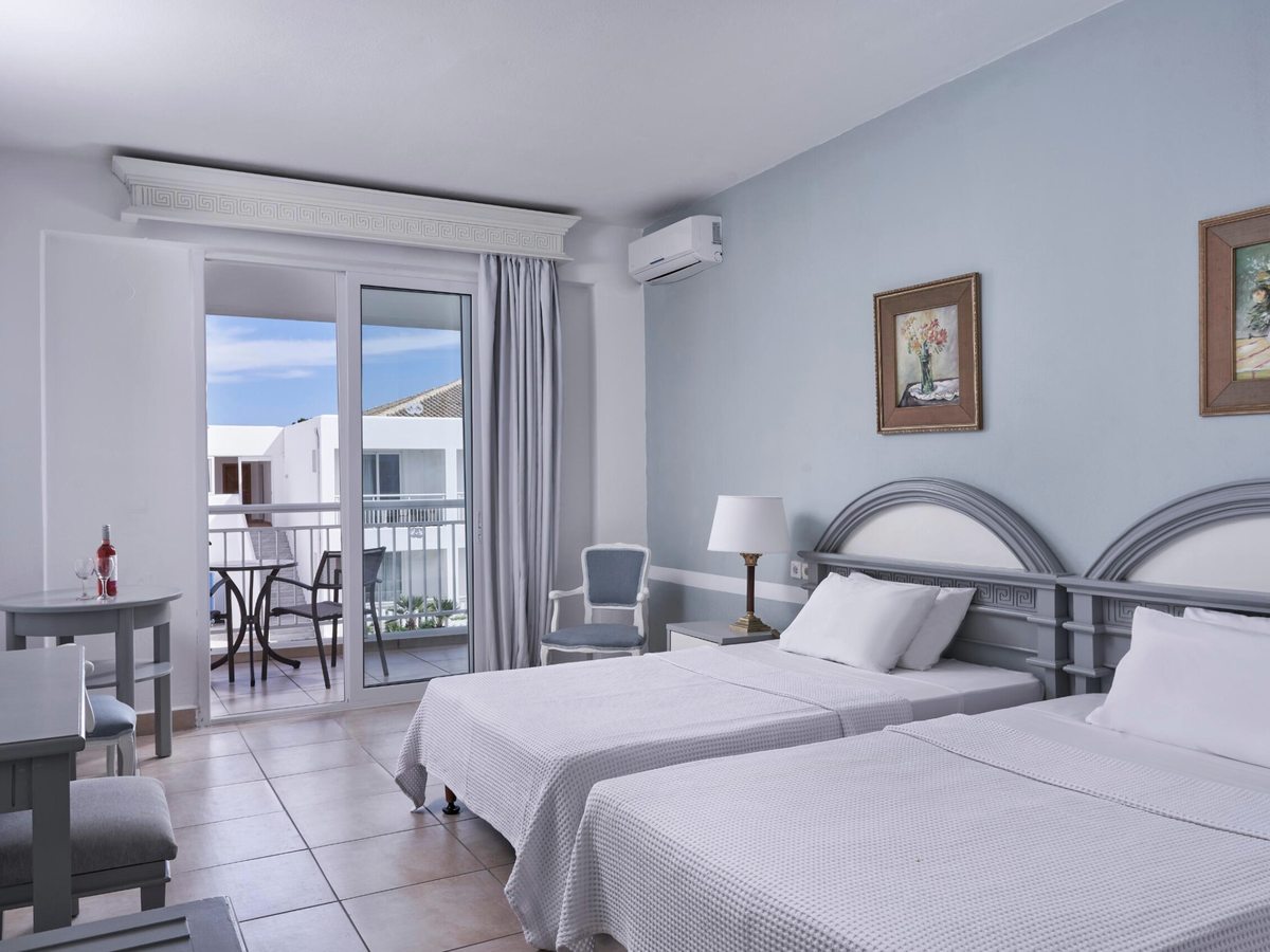 A pale blue twin room with two beds under grey arched headboards, framed pictures on the wall, a small table and chairs and sliding doors to a balcony.