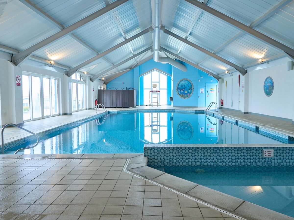 An indoor pool under a white pitched roof with skylights, a rectangular main pool with stainless handrails and a shallower section along one side.