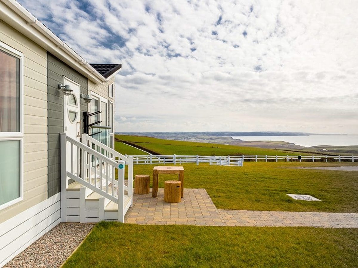 The end of a cream-clad holiday home with white steps up to its door and a paved patio with a wooden table and log stools, on grass behind a white fence.