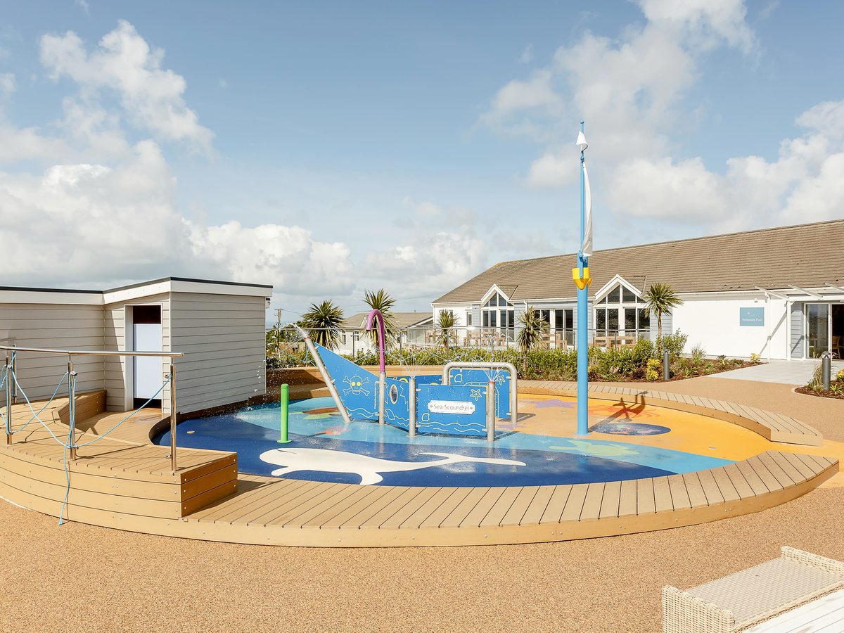 A circular splash area with a blue boat-shaped water feature, upright jets and a tall mast, ringed by timber decking, with the pool building behind.