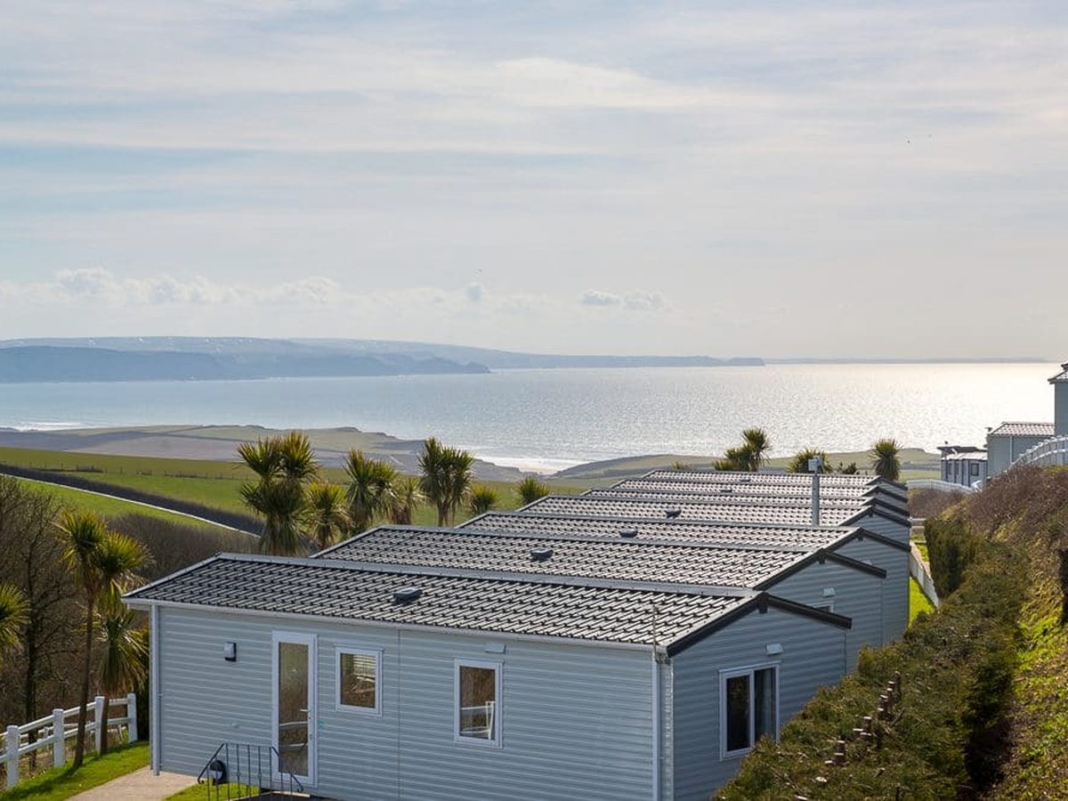 A grey holiday home on a grassy slope, with fields dropping away to a wide bay and a distant headland under a pale sky.