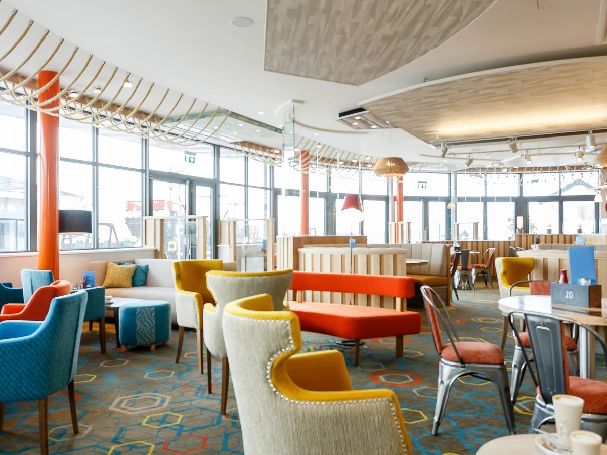 A lounge area with mustard and cream armchairs, an orange banquette and timber booth seating on a patterned blue carpet, beside full-height windows.