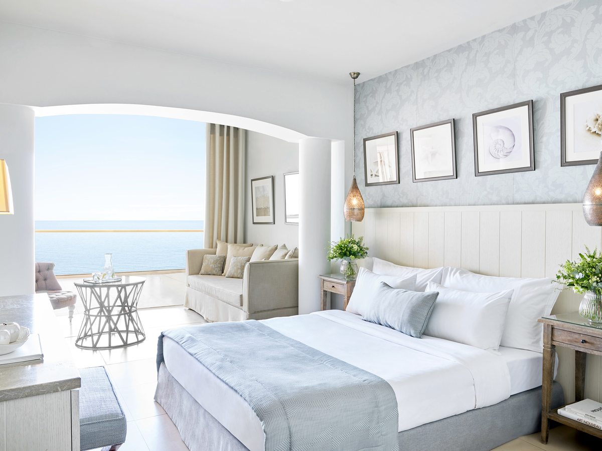 Bright room with a white panelled headboard, pale blue patterned wall and framed shell prints, opening through an arch to a sofa and a terrace above the sea.