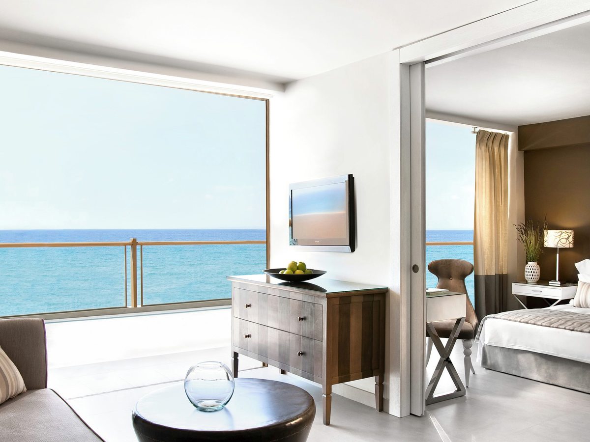 Suite living area with a grey sofa, wooden chest of drawers and wall-mounted television, a wide sea-facing terrace to the left and a bedroom through open sliding doors to the right.