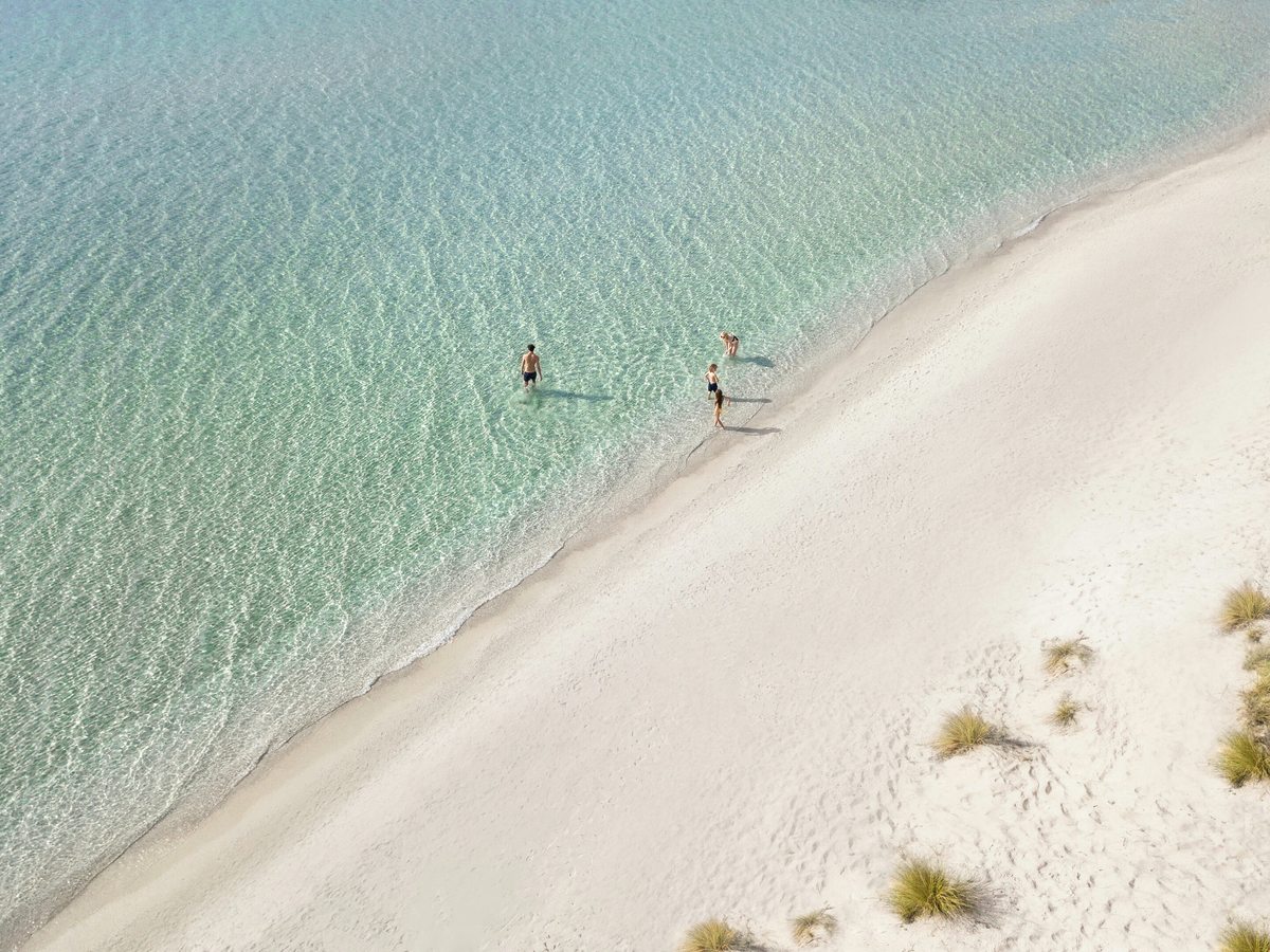 Aerial view of clear shallow water over pale sand with three people wading, beside a wide strip of white sand dotted with tufts of dune grass.