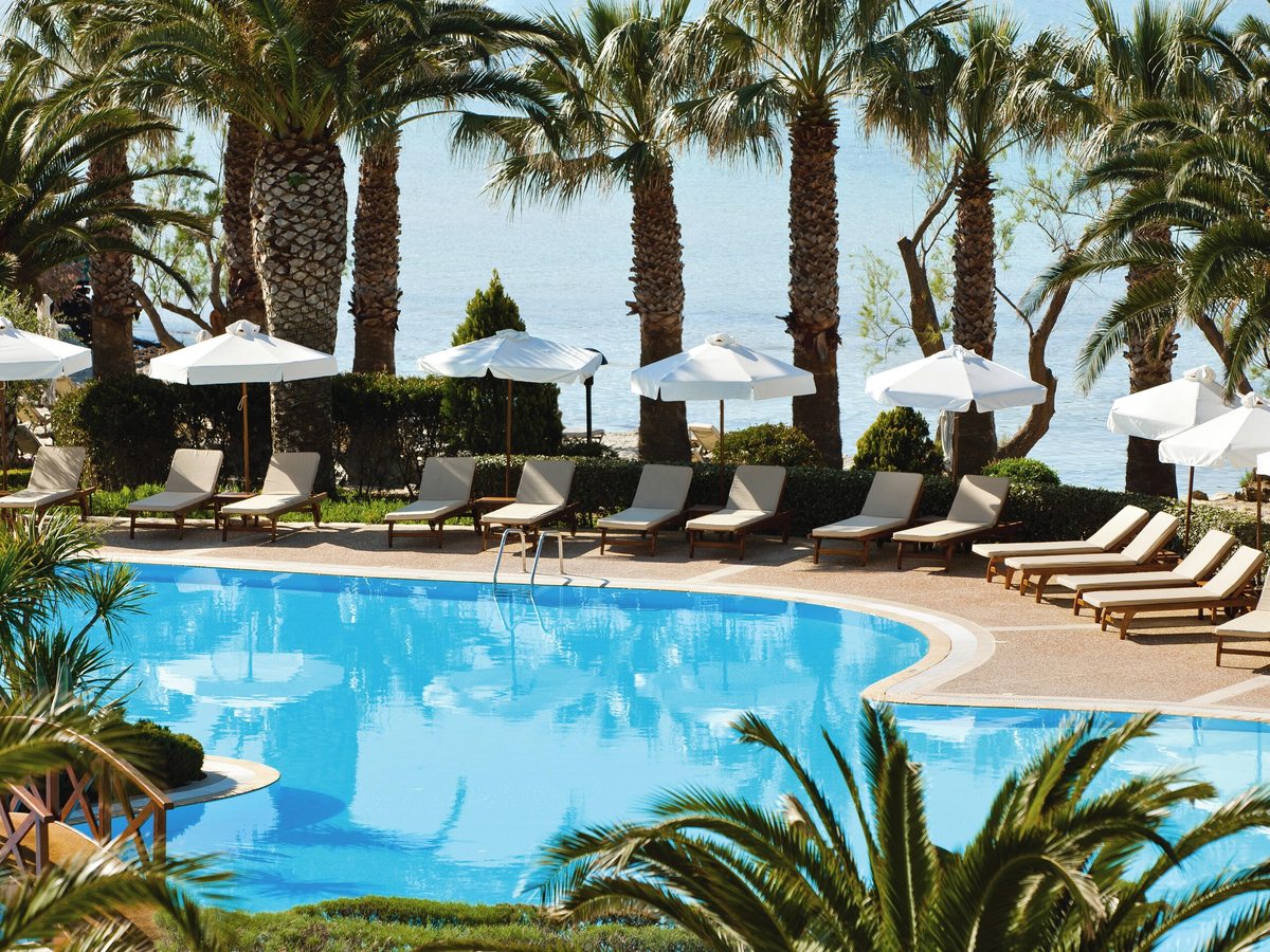 Curved turquoise pool edged by a wide terrace of wooden sun loungers and white parasols under tall palm trees, with the sea beyond.