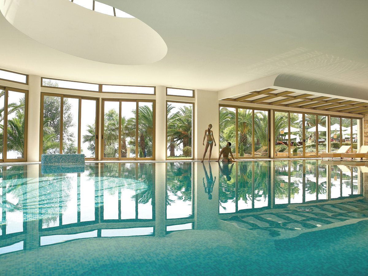 Indoor mosaic-tiled pool with floor-to-ceiling windows onto palms and gardens, a curved opening in the ceiling and two swimmers at the far end.