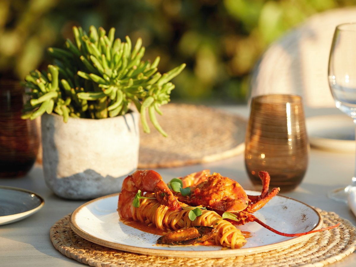 Half a grilled lobster served over pasta on a gold-rimmed plate, with a potted plant, wine glass and tumbler on the table behind.