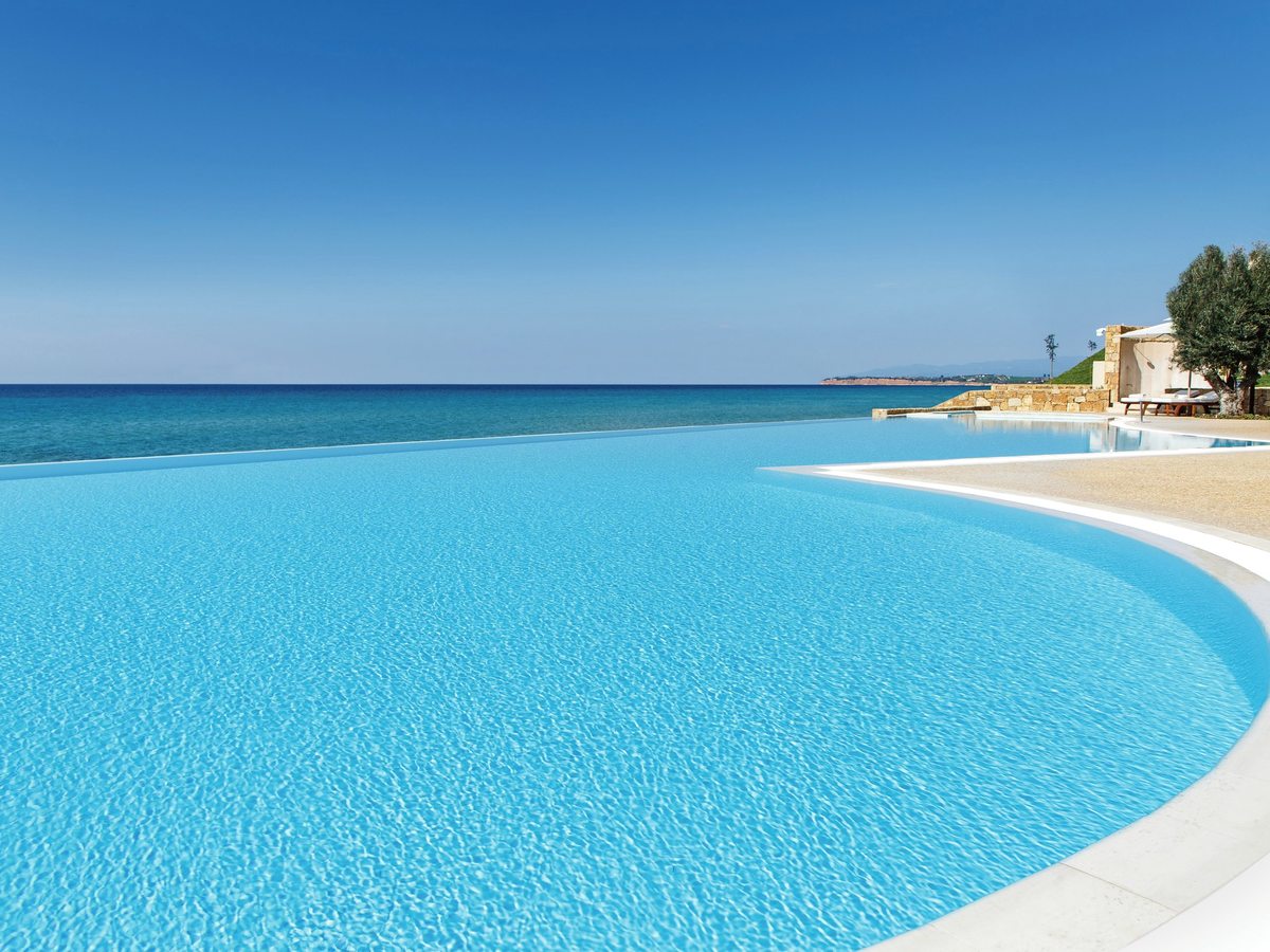 Wide curving infinity pool whose pale blue water meets the horizon of a calm sea, with a stone-walled cabana and an olive tree at the far end.