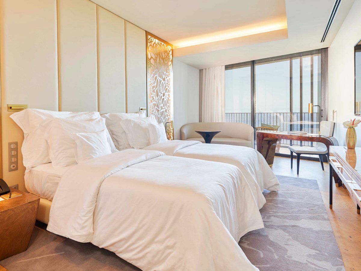 A bedroom with two single beds side by side, a cream padded headboard, a sofa and a desk beside floor-to-ceiling balcony doors.