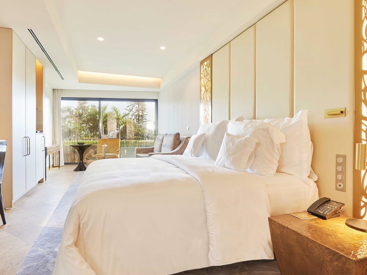 A bedroom with a large bed, fitted cream wardrobes and sliding doors onto a balcony with palms and garden planting beyond.