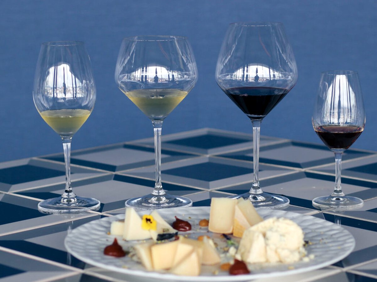 Four glasses of white and red wine on a blue-tiled table beside a plate of sliced cheeses, chutney and fruit.