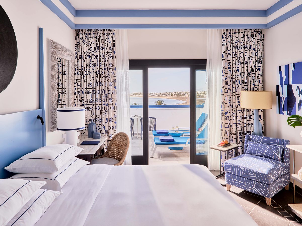 Double room with a blue upholstered headboard and patterned curtains, and glass doors opening onto a terrace with the sea beyond.