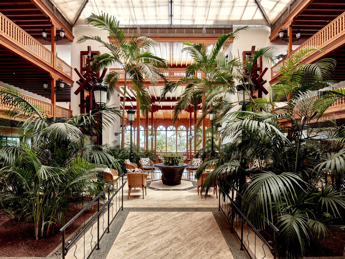 Hotel lobby atrium filled with tall palms and tropical planting under a glazed roof, with wooden balconies running round the upper floors.