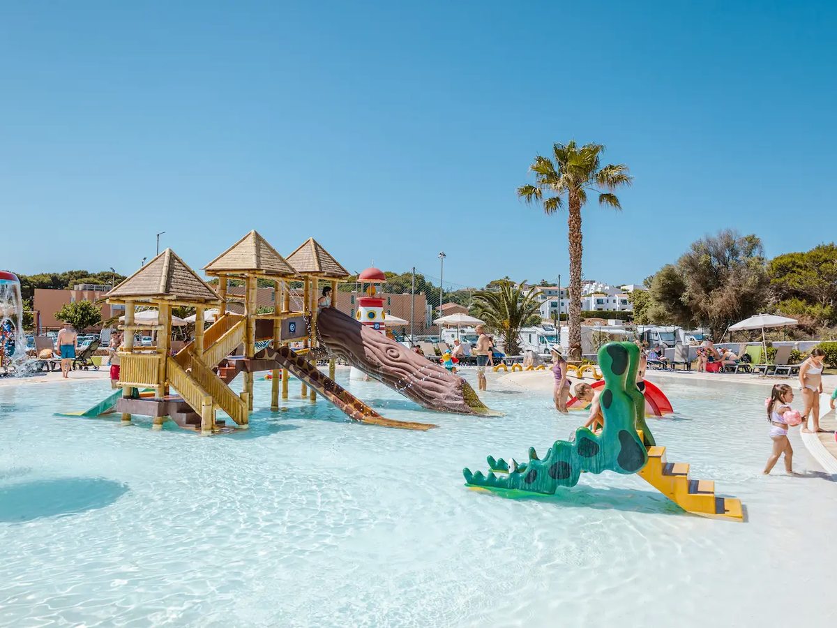 Children playing in a shallow pool with wooden play towers, a curling slide and a green dragon climbing feature.