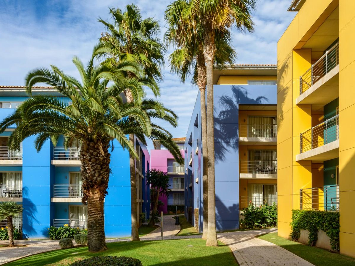 Four-storey blocks painted blue, yellow and pink around a lawn planted with palms, each floor with a run of balconies.