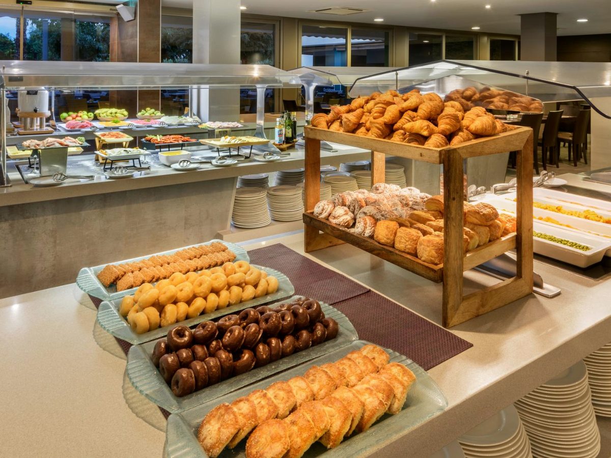 Trays of croissants, doughnuts and pastries on a buffet counter, with stacked plates and a chilled display of cold dishes behind.
