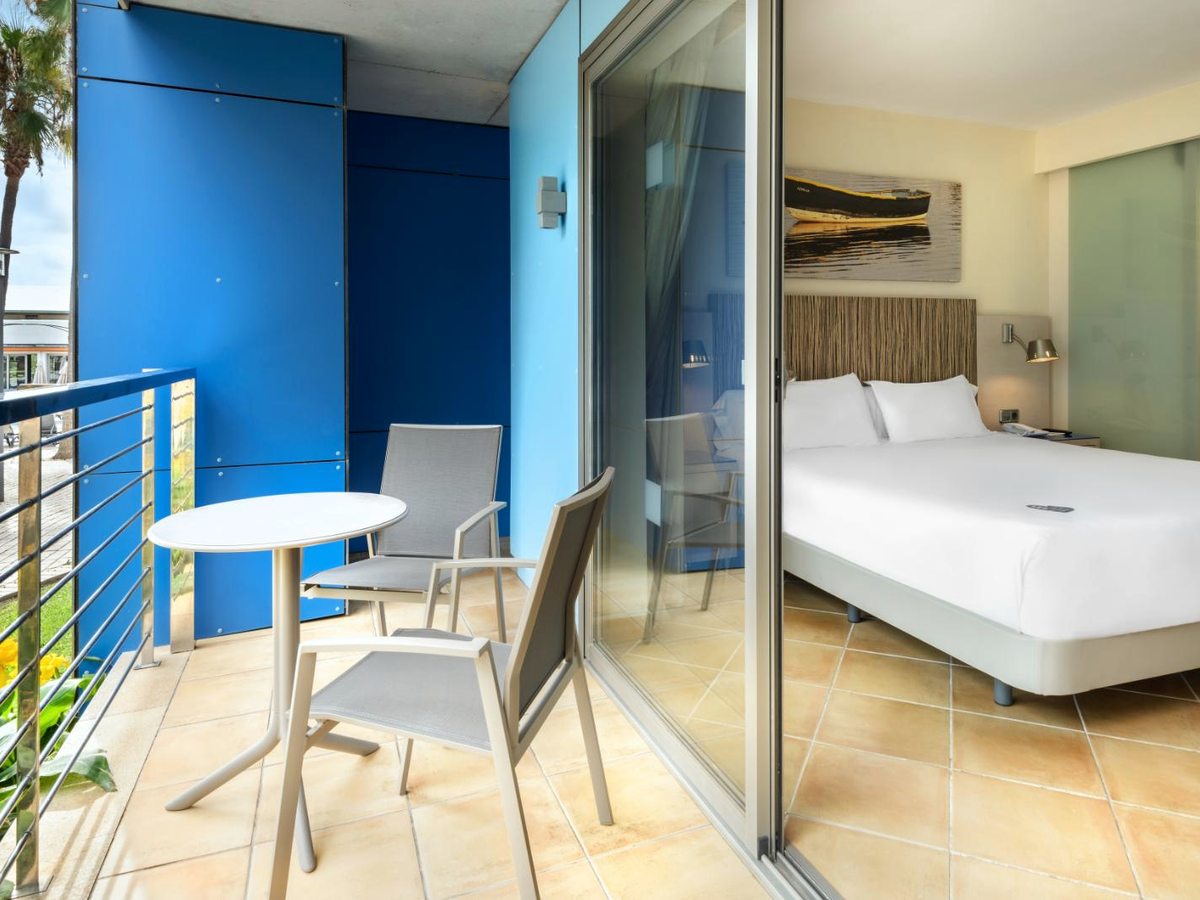 A balcony with a small round table and two chairs, a blue-painted wall alongside and sliding doors back into the bedroom.