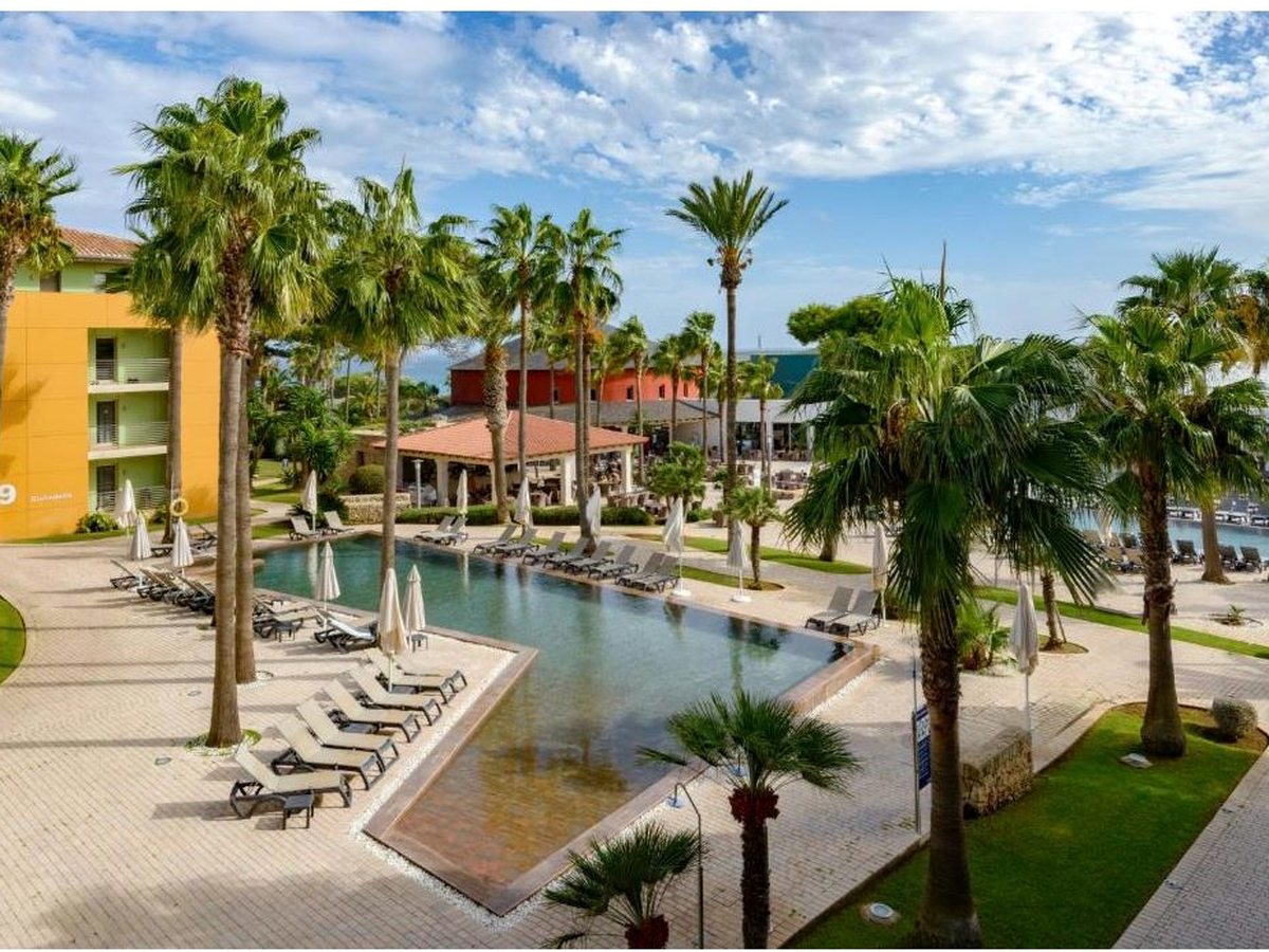 A large pool curving around planted islands of palms, with sun loungers along the paving and lawns and resort buildings behind.