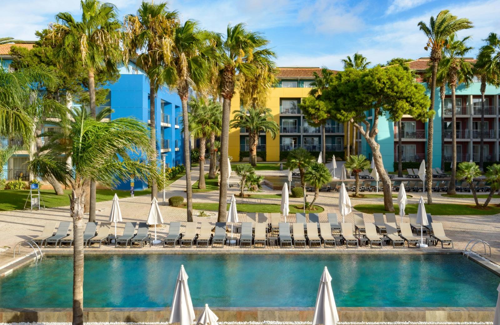 A long turquoise pool with a row of sun loungers and closed parasols along it, palms and lawns behind, and blue, yellow and green four-storey blocks beyond.