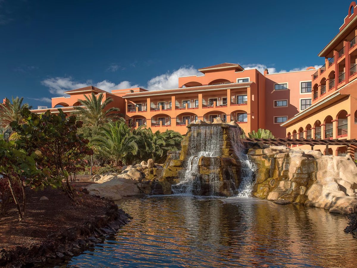 Terracotta hotel buildings with arched balconies rising behind a rock waterfall that falls into a garden pool, with palms and planting around it.