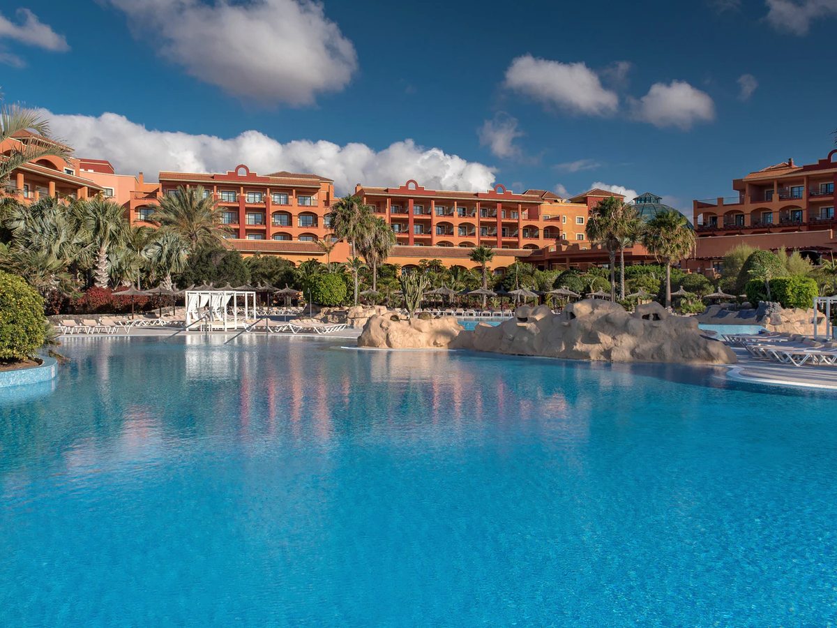 A large lagoon-shaped pool with rock edging and a shaded four-poster daybed, the terracotta resort buildings and palms behind it.