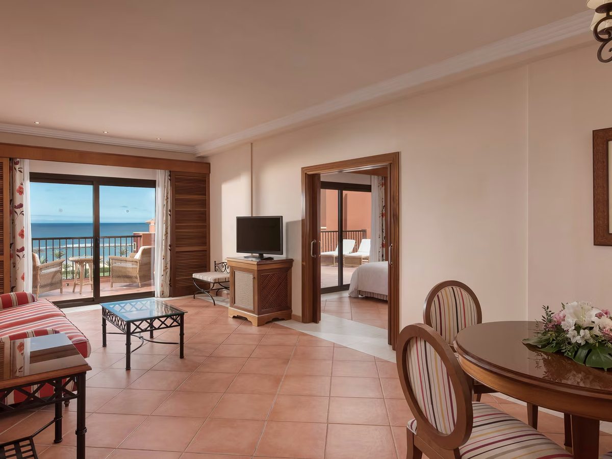 A suite living room with a sofa, coffee table and round dining table, the bedroom through open double doors and a sea-view terrace beyond.