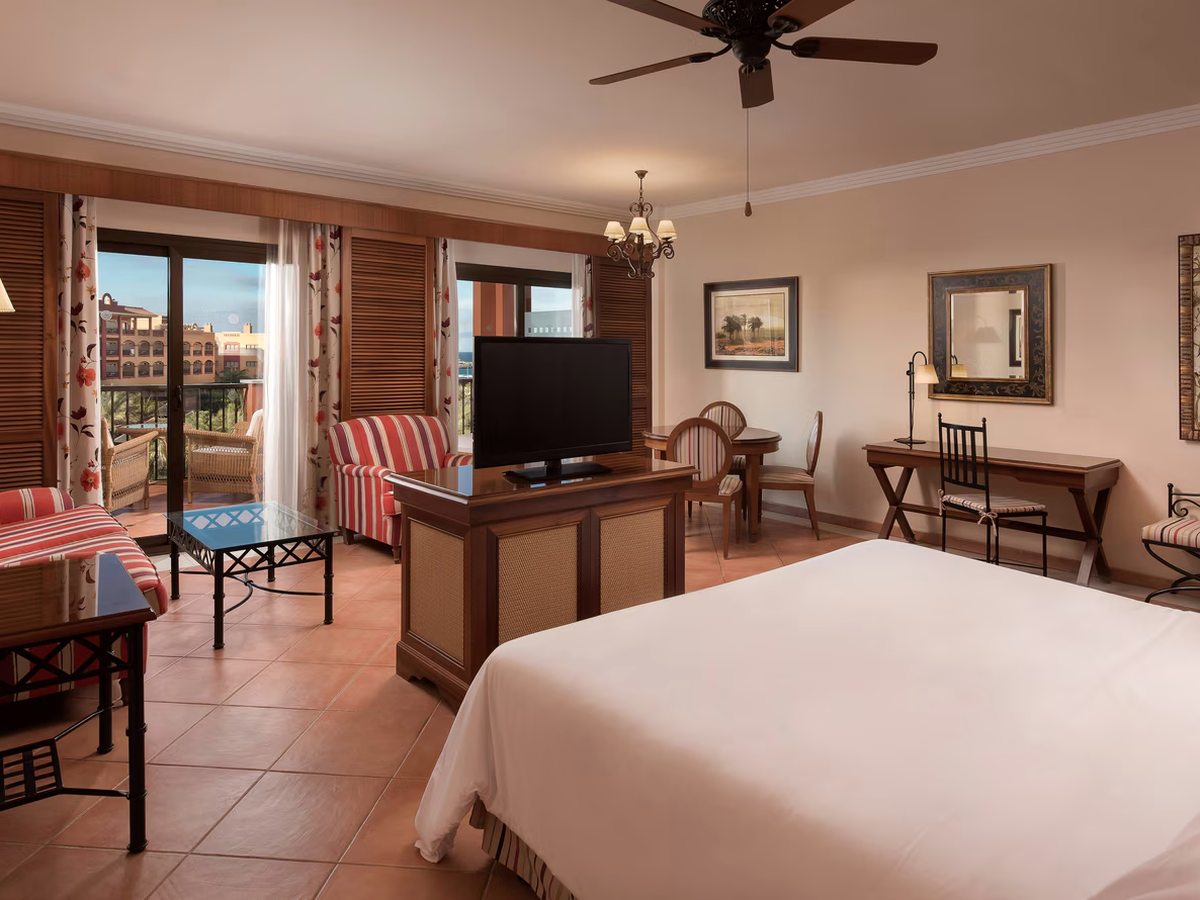 A guest room with a large bed, a striped sofa and armchairs, a writing desk and terracotta floor tiles, with shuttered doors onto a terrace.