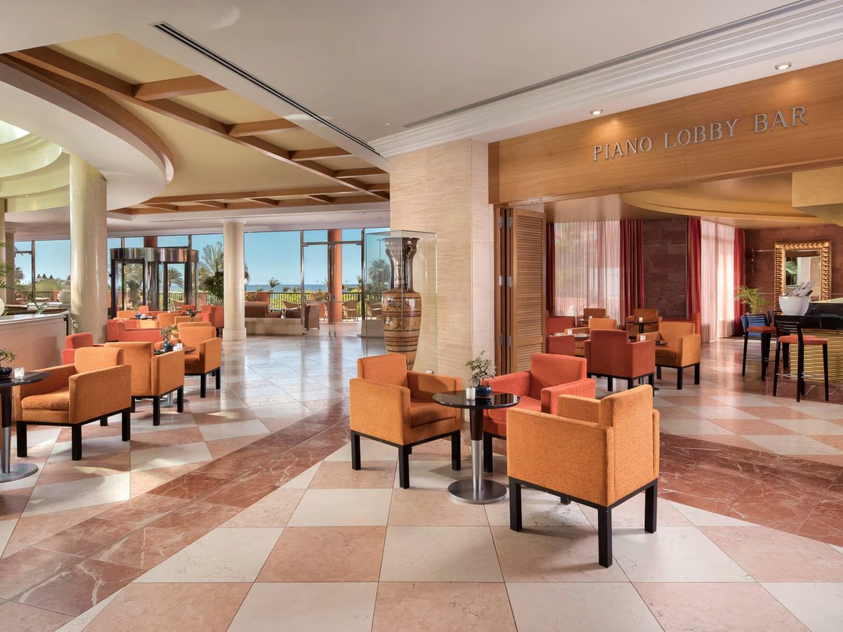 A marble-floored lobby lounge with orange armchairs and small round tables, the Piano Lobby Bar sign above its entrance and glass doors onto a sea view.