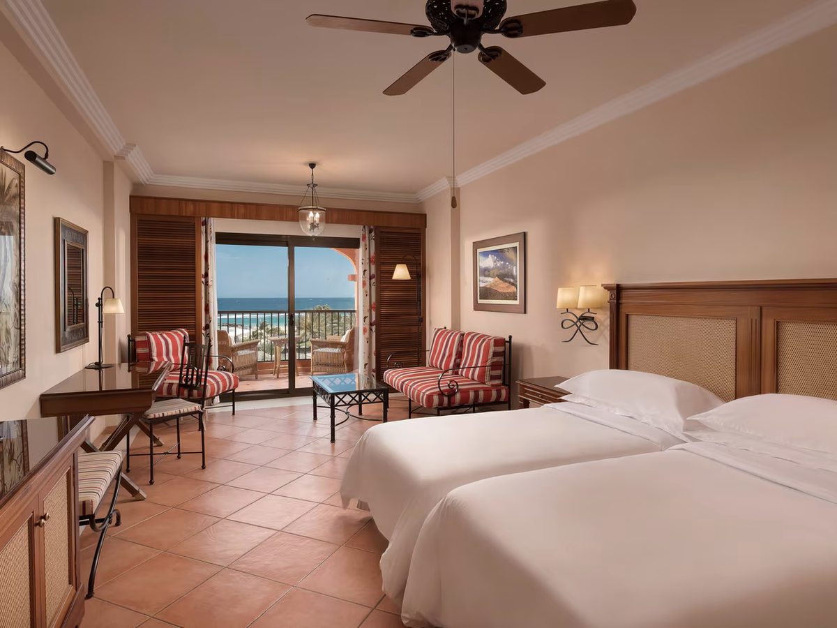 A twin room with two single beds side by side, a striped sofa and armchair, tiled floor and glass doors onto a balcony above the beach.